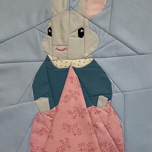 Puede incluir: Una pieza de arte textil con un diseño geométrico de conejo. El conejo tiene una cara y orejas de color gris claro con el interior de las orejas rosa, lleva una chaqueta verde azulado y un vestido rosa con un estampado floral. El fondo es azul claro.