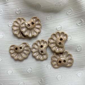 May include: Six beige ceramic buttons are displayed. Each button features a scalloped, flower-like design with two holes for sewing. The buttons are arranged on a white, textured fabric.