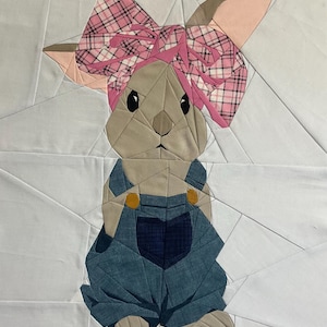 Puede incluir: Una obra de arte textil que representa un conejito con un peto vaquero y un lazo rosa a cuadros. El conejito tiene pelaje marrón claro, ojos negros y orejas internas rosas. El fondo es blanco con patrones geométricos.