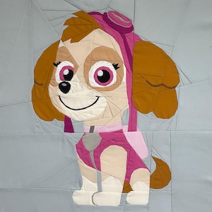 Op de afbeelding: Een kleurrijk stoffen kunstwerk met een cartoonhond, voornamelijk in roze, bruine en beige tinten. De hond heeft grote roze ogen, een glimlach en draagt een roze pilotenhelm. De achtergrond is een lichtblauw patchwork ontwerp.