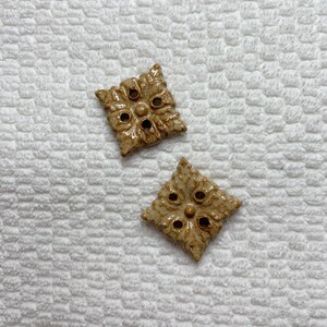 Peut inclure: Deux boutons décoratifs en céramique avec un motif floral. Les boutons sont de forme carrée avec un glaçage brun clair et des accents brun foncé. Les boutons ont un motif floral en relief et de petits trous.