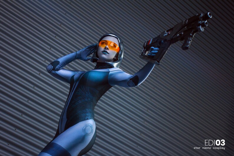 May include: A person in a futuristic blue and black bodysuit with orange-tinted goggles and a silver helmet, holding a black and grey weapon. The image is set against a corrugated metal background. The text "EDI03" and "star nacho cosplay" are visible.