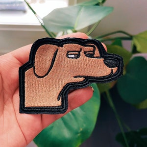 May include: An embroidered patch of a dog with a sad expression. The dog is brown with a black outline and is looking to the left.