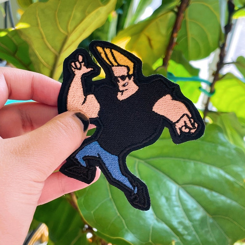 May include: An embroidered patch featuring a cartoon character with blonde hair, wearing a black shirt and blue jeans. The character is running with one arm raised.