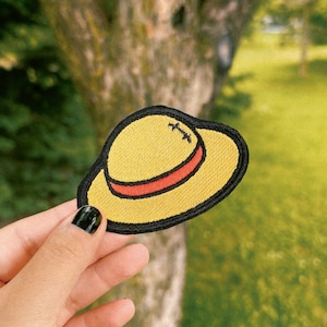 Op de afbeelding: Een geborduurde patch van een strohoed met een rode band, in geel en zwart.