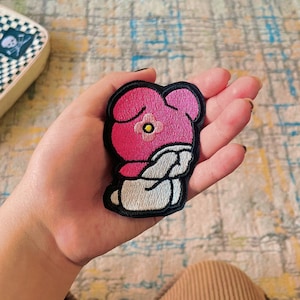 May include: Pink and white embroidered iron-on patch of a cartoon character with a flower on its head. The character is wearing a white hooded garment and has its head down.