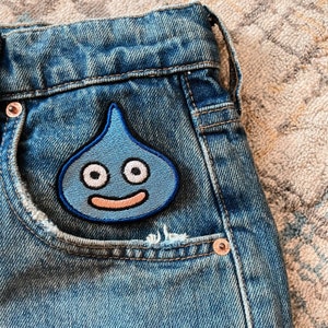 Könnte beinhalten: Blaue Jeans aus Denim mit einem blauen und weißen gestickten Aufnäher einer lächelnden, tropfenförmigen Kreatur mit zwei großen Augen.