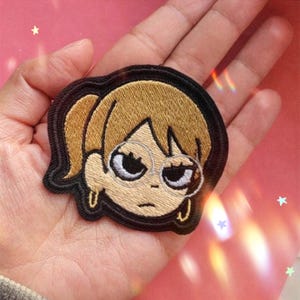 May include: Embroidered patch featuring a cartoon character with blonde hair in a ponytail, wearing glasses and earrings. The patch has a black border and is set against a pink background with star-shaped sparkles.