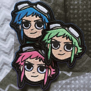 Puede incluir: Tres parches bordados con personajes de dibujos animados con ojos grandes y cabello de colores. Los personajes llevan gafas protectoras y tienen una expresión sonriente. Los parches están hechos de fieltro y tienen un contorno negro.