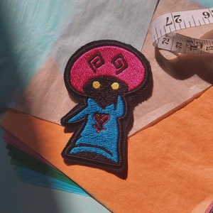 May include: Embroidered patch of a cartoon character with a pink mushroom hat, blue shirt, and black body. The character has yellow eyes and a red heart with a black outline on its chest.