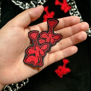 May include: Three red embroidered flower patches with black outlines. The patches are on a hand, with a black background.