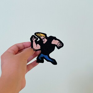 May include: An embroidered patch featuring a cartoon character running. The character has blonde hair and is wearing a black shirt and blue trousers.