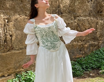 May include: A white dress with a green floral print bodice. The dress has a fitted bodice and a flowing skirt. The model is standing in front of a stone wall.