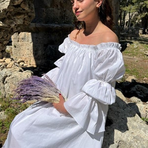 May include: A woman in a white off-the-shoulder dress with ruffled sleeves holds a bouquet of dried lavender.