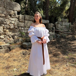 May include: A white off-the-shoulder dress with long, puffy sleeves and a brown belt. The dress is worn by a person standing in a forest setting.