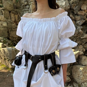 May include: A white off-the-shoulder peasant top with long sleeves and a black leather belt with decorative metal rings and pouches.