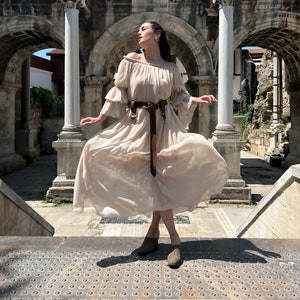 K&ouml;nnte beinhalten: Ein flie&szlig;endes, schulterfreies beiges Maxikleid mit gestuften, ger&uuml;schten &Auml;rmeln und gerafftem Ausschnitt. Das Kleid wird in der Taille mit einem breiten, dunkelbraunen Lederg&uuml;rtel mit gro&szlig;er runder Schnalle und zwei herabh&auml;ngenden Riemen gerafft.