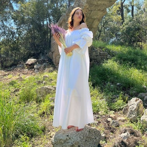 May include: A woman in a white off-the-shoulder dress stands on a rock in a forest setting. She is holding a bouquet of purple flowers.