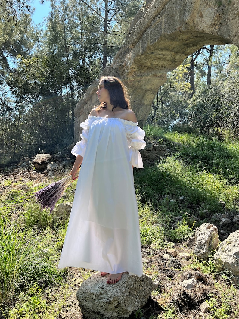 May include: A woman wearing a white off-the-shoulder maxi dress with puffed sleeves stands on a rock in a forest setting. She is holding a bouquet of dried lavender.