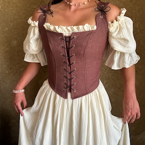 May include: A brown corset with a lace-up front and a ruffled neckline. The corset is worn over a white off-the-shoulder blouse and a white skirt.