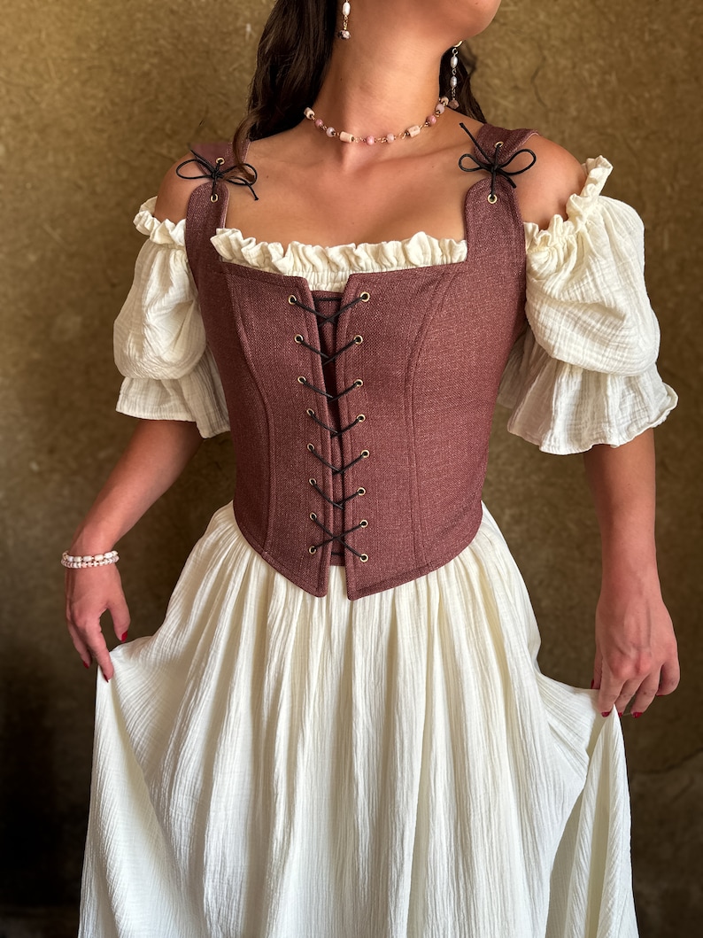 May include: A brown corset with a lace-up front and a ruffled neckline. The corset is worn over a white off-the-shoulder blouse and a white skirt.