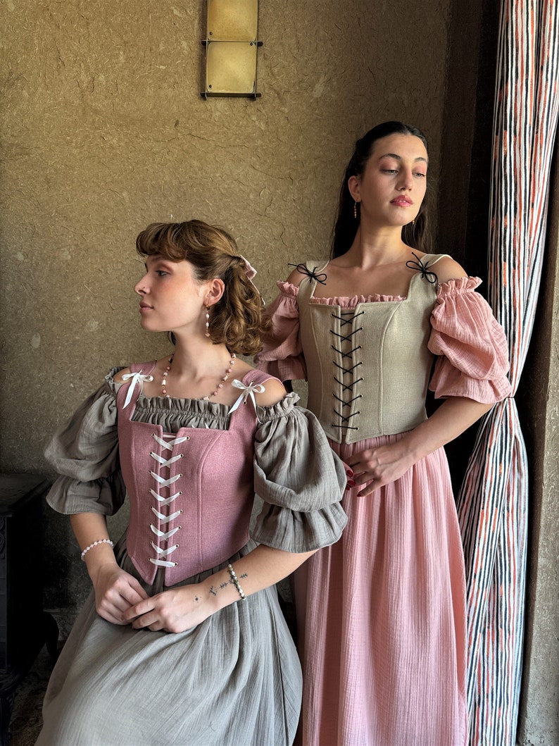 May include: Two women wearing off-the-shoulder tops with lace-up corsets. The woman on the left is wearing a pink corset and a gray skirt. The woman on the right is wearing a beige corset and a pink skirt.