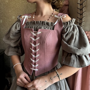May include: A pink linen corset with white lacing and gold grommets. The corset has a square neckline and is worn over a brown linen top with puffed sleeves.