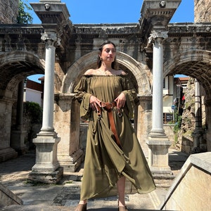 K&ouml;nnte beinhalten: Ein olivgr&uuml;nes schulterfreies Maxikleid mit langen, weiten &Auml;rmeln und gerafftem Ausschnitt. Das Kleid wird in der Taille mit einem breiten braunen Lederg&uuml;rtel mit silberfarbener Schnalle zusammengehalten. Der Stoff wirkt leicht und flie&szlig;end.
