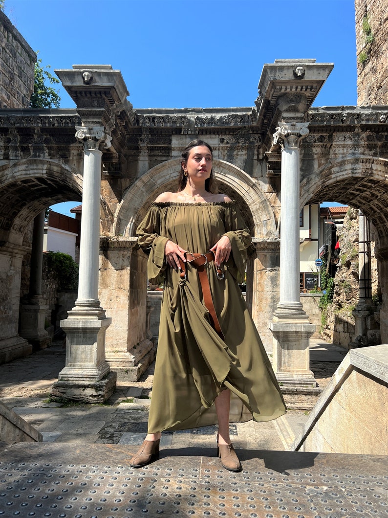 K&ouml;nnte beinhalten: Ein olivgr&uuml;nes schulterfreies Maxikleid mit langen, weiten &Auml;rmeln und gerafftem Ausschnitt. Das Kleid wird in der Taille mit einem breiten braunen Lederg&uuml;rtel mit silberfarbener Schnalle zusammengehalten. Der Stoff wirkt leicht und flie&szlig;end.