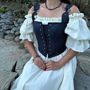 May include: A blue denim corset with a white lace trim and gold eyelets. The corset is worn over a white dress with puffed sleeves.