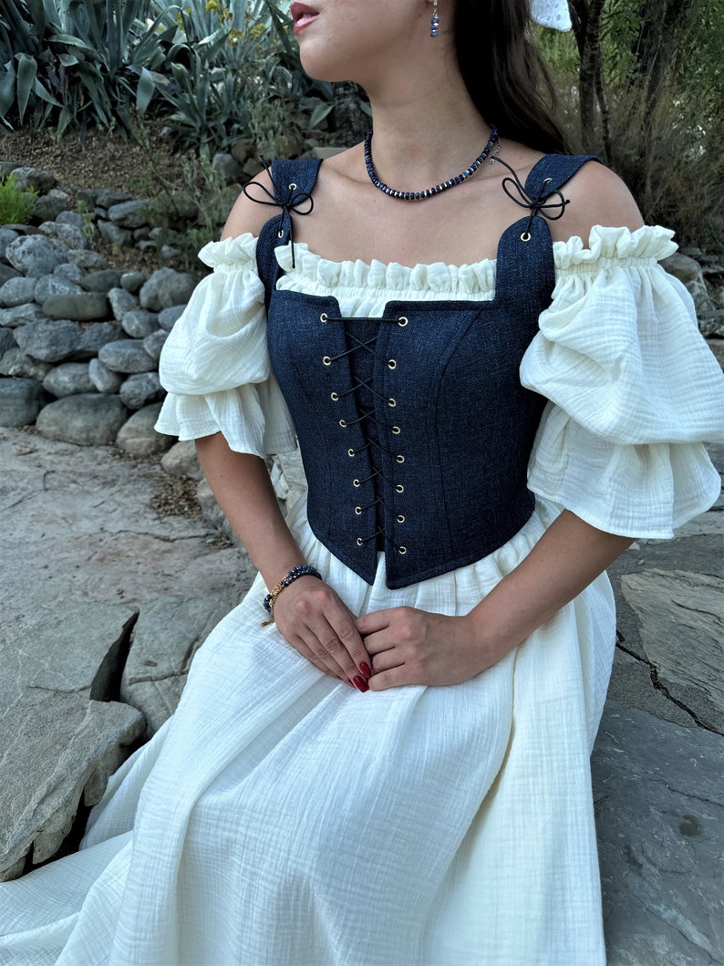 May include: A blue denim corset with a white lace trim and gold eyelets. The corset is worn over a white dress with puffed sleeves.