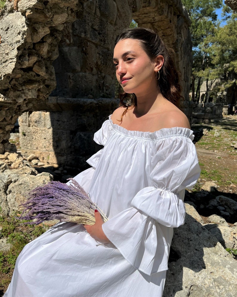 May include: A woman in a white off-the-shoulder dress with ruffled sleeves holds a bouquet of dried lavender.