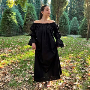 May include: A black off-the-shoulder maxi dress with long, puffy sleeves. The dress is made of a soft, flowing fabric and has a relaxed fit. The model is standing in a grassy area with fallen leaves.
