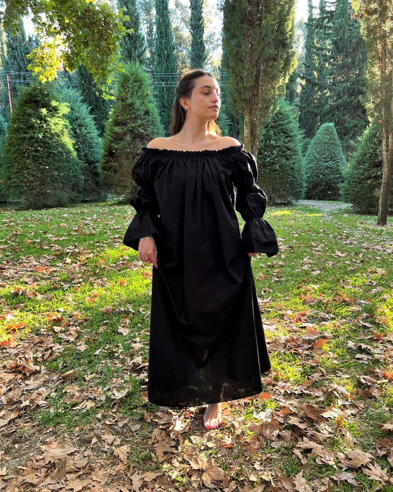 May include: A black off-the-shoulder maxi dress with long, puffy sleeves. The dress is made of a soft, flowing fabric and has a relaxed fit. The model is standing in a grassy area with fallen leaves.