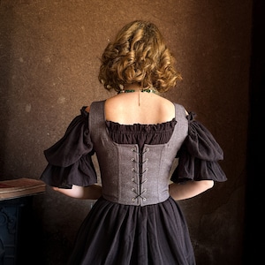 May include: A woman wearing a brown and black dress with a gray corset. The corset has a lace-up back and is cinched at the waist. The woman is sitting on a wooden stool.