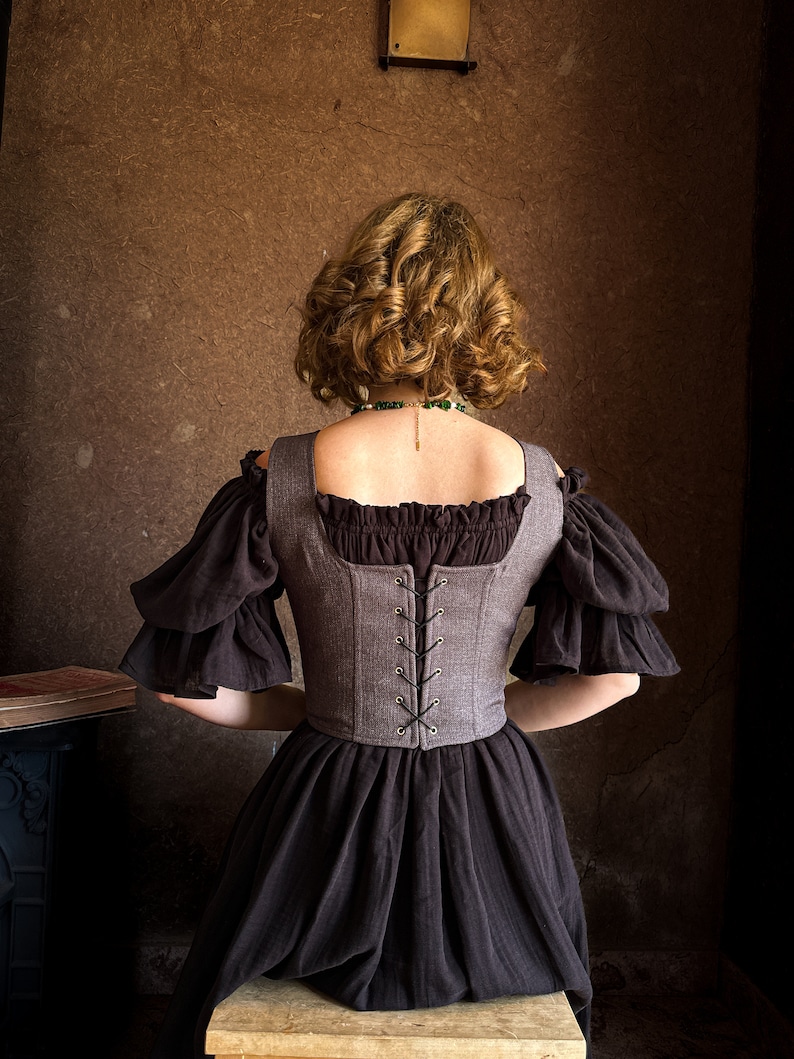 May include: A woman wearing a brown and black dress with a gray corset. The corset has a lace-up back and is cinched at the waist. The woman is sitting on a wooden stool.