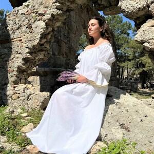 May include: A woman in a white off-the-shoulder dress sits on a rock in front of a stone archway. She is holding a bouquet of purple flowers.