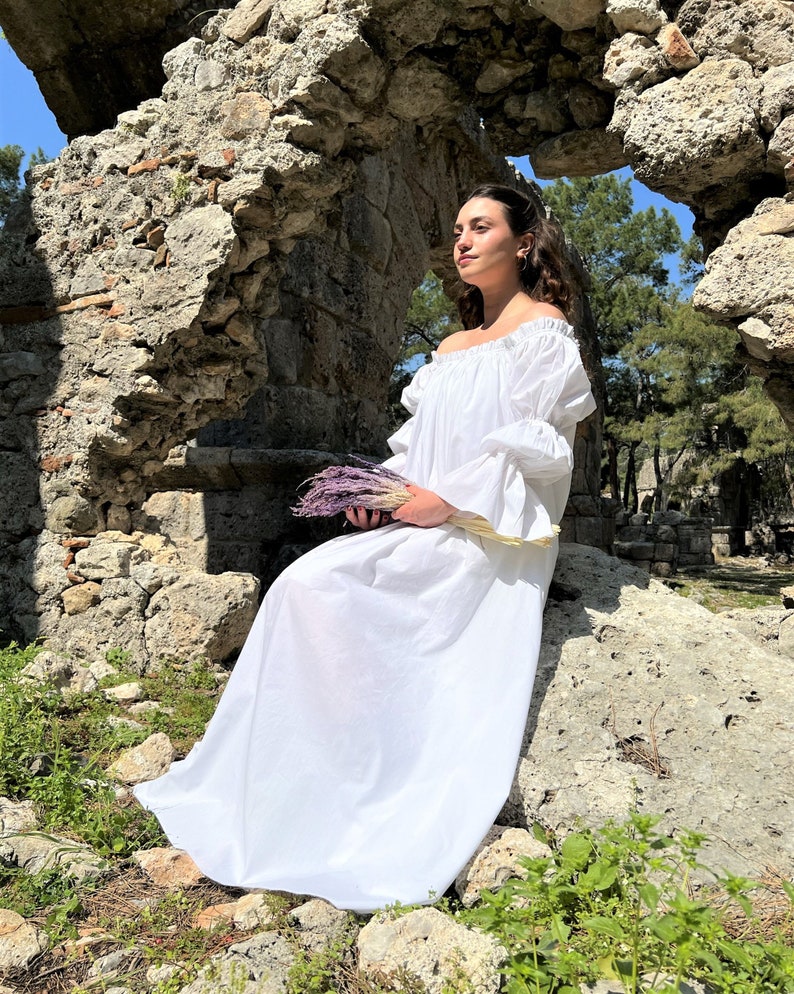 May include: A woman in a white off-the-shoulder dress sits on a rock in front of a stone archway. She is holding a bouquet of purple flowers.
