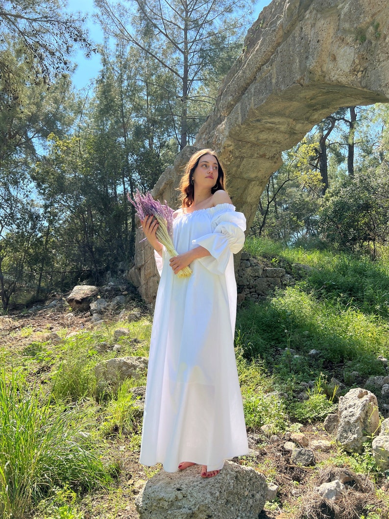 May include: A woman in a white off-the-shoulder dress stands on a rock in a forest setting. She is holding a bouquet of purple flowers.