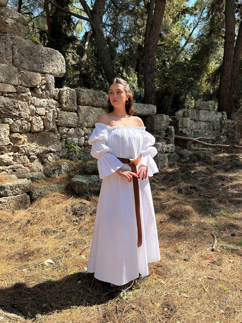 May include: A white off-the-shoulder dress with long, puffy sleeves and a brown belt. The dress is worn by a person standing in a forest setting.