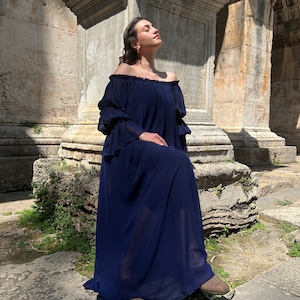 K&ouml;nnte beinhalten: Ein bodenlanges, marineblaues Kleid mit schulterfreiem Ausschnitt und gerafftem Gummizug, langen, gestuften R&uuml;schen&auml;rmeln und einer locker fallenden Silhouette aus leichtem, halbtransparentem Stoff, der bis zum Boden reicht.