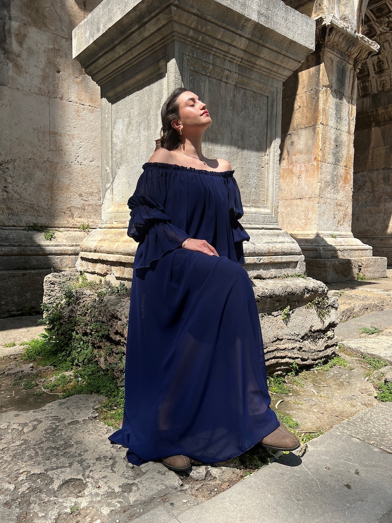 K&ouml;nnte beinhalten: Ein bodenlanges, marineblaues Kleid mit schulterfreiem Ausschnitt und gerafftem Gummizug, langen, gestuften R&uuml;schen&auml;rmeln und einer locker fallenden Silhouette aus leichtem, halbtransparentem Stoff, der bis zum Boden reicht.