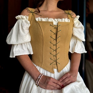 May include: A woman wearing a white dress with a mustard yellow linen corset with green lacing. The corset has a square neckline and is tied at the front. The woman is wearing a beaded bracelet on her wrist.