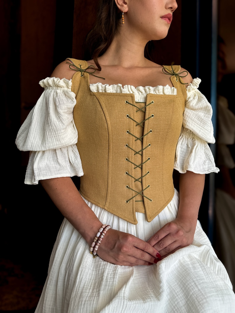 May include: A woman wearing a white dress with a mustard yellow linen corset with green lacing. The corset has a square neckline and is tied at the front. The woman is wearing a beaded bracelet on her wrist.