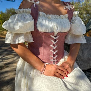 May include: A pink linen corset with white lace-up detailing and straps. The corset is worn over a white off-the-shoulder blouse with ruffled sleeves. The model is wearing a white skirt and a pink and white beaded bracelet.