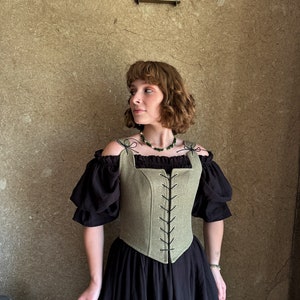 May include: A green linen corset with black lacing and a square neckline. The corset is worn over a black off-the-shoulder top and a black skirt.