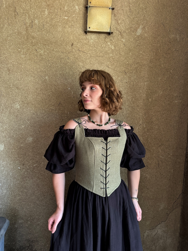 May include: A green linen corset with black lacing and a square neckline. The corset is worn over a black off-the-shoulder top and a black skirt.