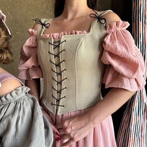 May include: A beige linen corset with black lacing and gold grommets. The corset has a square neckline and is worn over a pink dress with puffed sleeves.