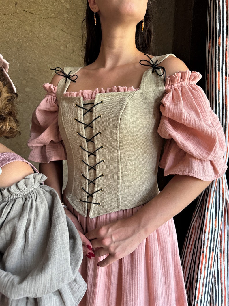 May include: A beige linen corset with black lacing and gold grommets. The corset has a square neckline and is worn over a pink dress with puffed sleeves.