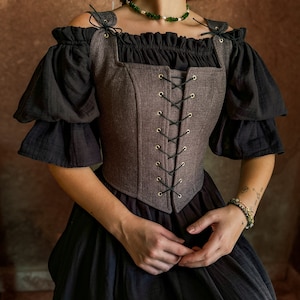 May include: A brown linen corset with black lacing and gold eyelets. The corset has a square neckline and is worn over a black off-the-shoulder top.